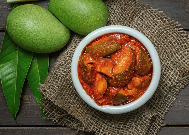 mango pickle or aam ka aachar or achar in a bowl on wooden background theme with raw mangos around that